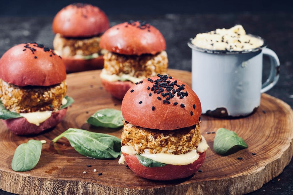 Beetroot bun sliders with crumbed patties, spinach and aioli on a wooden serving board.