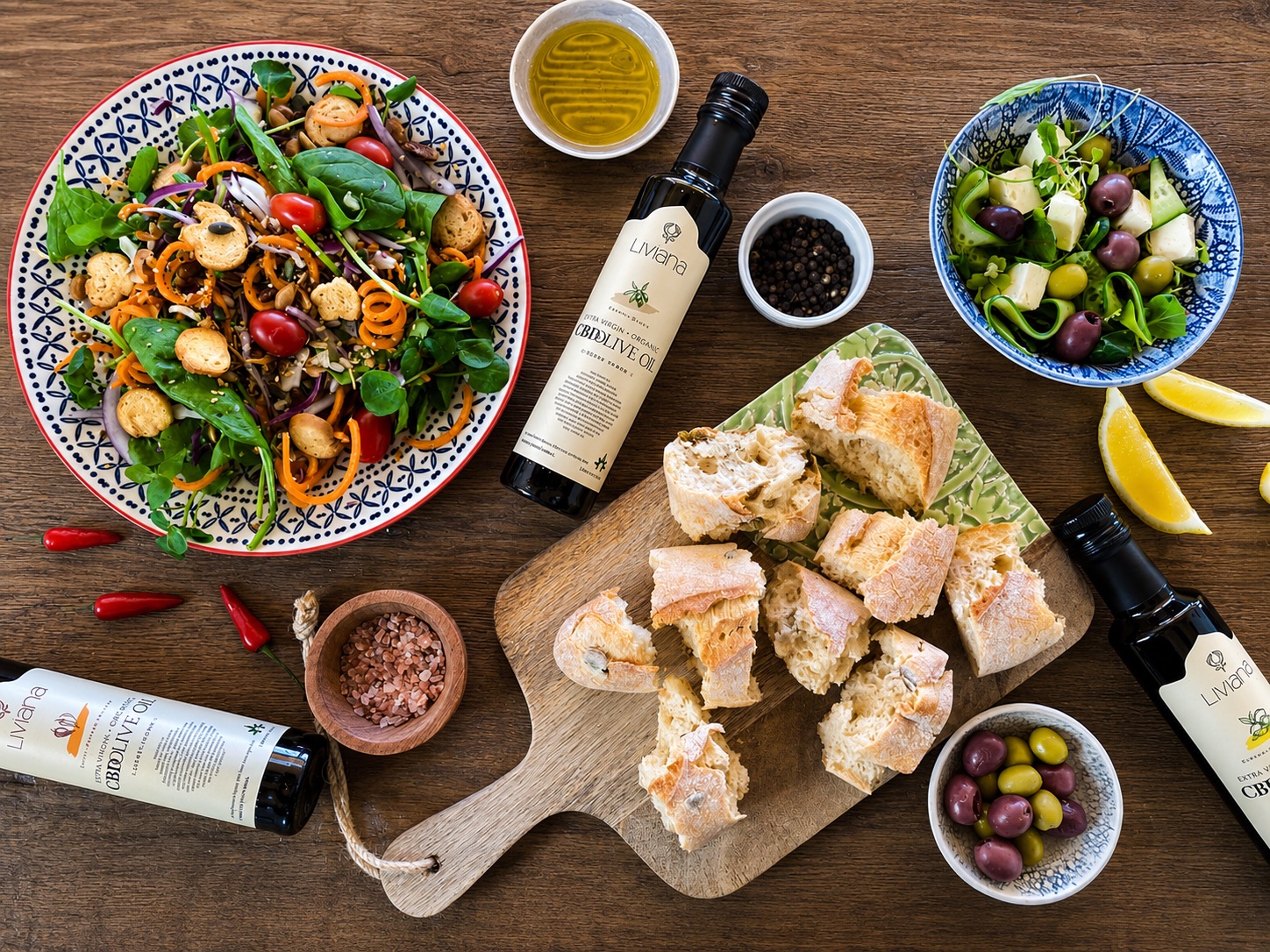 Overhead flat lay of Liviana extra virgin olive oil bottles with salads, ciabatta, olives and spices on a wooden table.