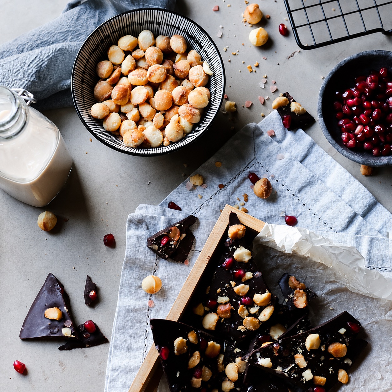 Dark chocolate macadamia bark with pomegranate, styled with a bowl of roasted macadamias and nut milk.