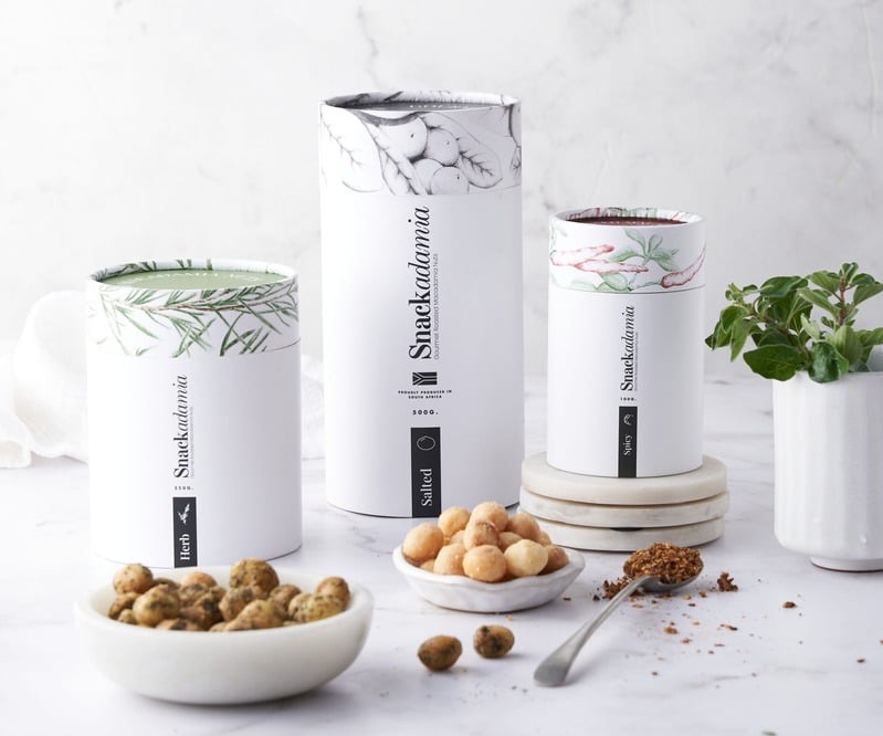 Snackadamia canisters in Herb, Salted and Spicy variants with bowls of roasted macadamias.